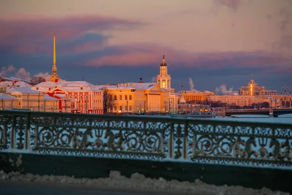 Winter panoramisch uitzicht op St. Petersburg bij zonsondergang, gouden torenspits van Peter en Paul fort, Kunstkamera toren, dijk met brug op de achtergrond, stoom over bevroren Neva rivier, lucht van oranje kleur — Stockfoto