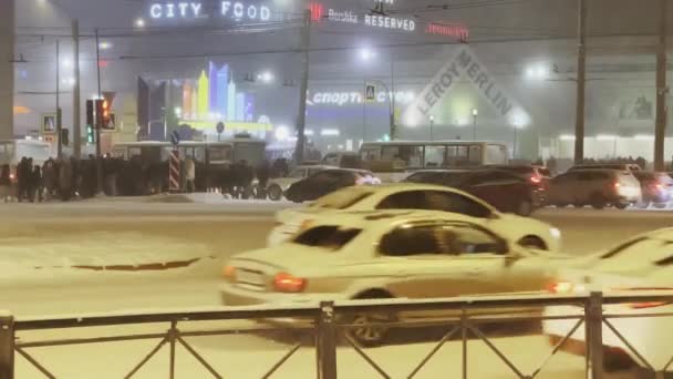 Ryssland, St Petersburg, 26 december 2021: Fotgängare som korsar korsningen, biltrafik, kraftigt snöfall i staden jäkt på natten, uge köpcentrum, snöstorm, flerfärgade skyltar på byggnaden — Stockvideo