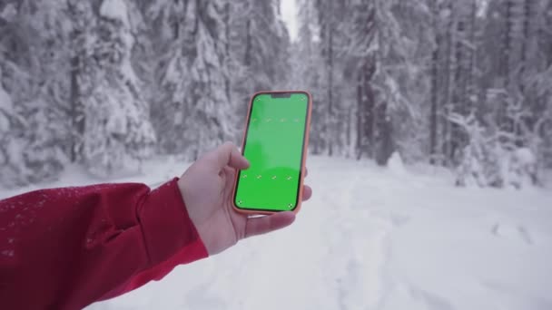The smartphone in a mans hand, snowy forest in the background, fingers move on the green screen of the phone, finger moves the screen to the right, a man gets lost and is looking for a way — Wideo stockowe