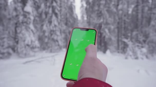 The smartphone in a mans hand, snowy forest in the background, fingers move on the green screen of the phone, finger moves the screen to the right, a man gets lost and is looking for a way — Video Stock