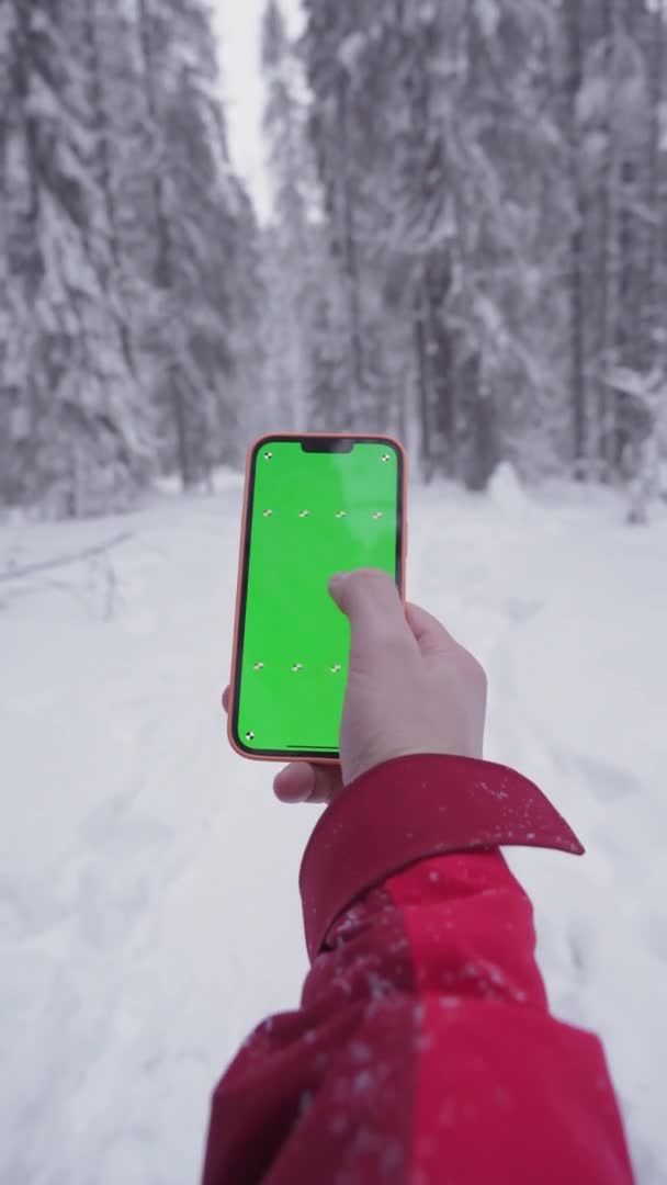 The smartphone in a mans hand, snowy forest in the background, fingers move on the green screen of the phone, finger moves the screen to the right, a man gets lost and is looking for a way — Wideo stockowe