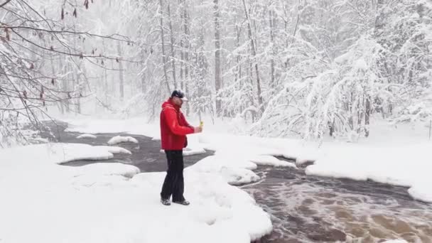 写真家は、スマートフォンで雪の嵐で野生の自然を写真やビデオに行い、彼は赤い色のジャケット、冬の木材、氷、雪に覆われた木の野生の凍結小さな川に身を包んだ — ストック動画