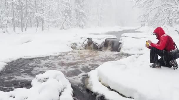 The photographer does to a photo and video the wild nature at snow storm by smartphone, he is dressed in red color jacket, the wild frozen small river in the winter wood, ice, snow-covered trees — Wideo stockowe