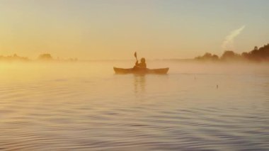 Kayaker floats on calm water through the morning fog above the water at sunrise, the silhouette of a man with a paddle on a kayak, the golden color of the water, warm water and cold air, magic light
