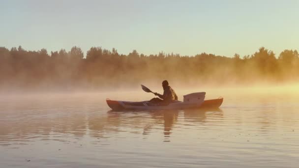 Russia, Karelia, 06 July 2021: Kayaker floats on calm water through the morning fog above the water at sunrise, the silhouette of a man with a paddle on a kayak, the golden color, magic light — стокове відео