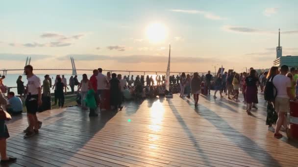 Rusia, San Petersburgo, 18 de agosto de 2019: La gente se relaja en el terraplén del complejo de exposiciones Sevkabel en un cálido día de verano al atardecer, la gente está bailando, un puente de cable se quedó en el fondo — Vídeos de Stock