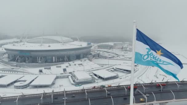Rusland, St. Petersburg, 22 januari 2022: De drone vliegt in de buurt van het nieuwe stadion Gazprom Arena in een sneeuwstorm, de vlag van het Zenit team zwaait op een enorme vlaggenmast, veel auto 's op de snelweg — Stockvideo