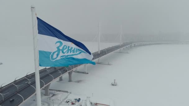 Russie, Saint-Pétersbourg, 22 janvier 2022 : Drapeau de l'équipe de football Zenit se balance sur un énorme mât de drapeau dans une tempête de neige, drone vole le long du pont à haubans près du nouveau stade Gazprom Arena — Video