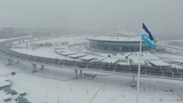 Ryssland, St Petersburg, 22 januari 2022: Drönaren flyger nära den nya arenan Gazprom Arena i en snöstorm, flaggan av Zenit laget sveper på en enorm flaggstång, en hel del bilar på motorvägen — Stockvideo