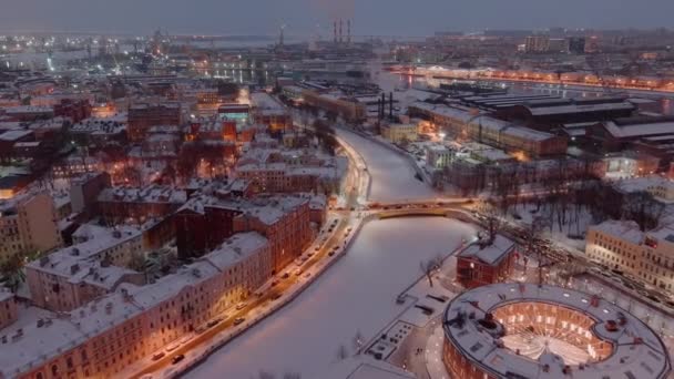 Luftaufnahme des New Holland Parks an einem verschneiten Winterabend, nächtliche Beleuchtung von Gebäuden und Straßen, nächtliches Stadtbild, Eisbahn im Freien, Hafenkräne von St. Petersburg im Hintergrund — Stockvideo