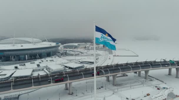 Russie, Saint-Pétersbourg, 22 janvier 2022 : Le drone vole près du nouveau stade Gazprom Arena dans une tempête de neige, le drapeau de l'équipe Zenit oscille sur un énorme mât de drapeau, beaucoup de voitures sur l'autoroute — Video