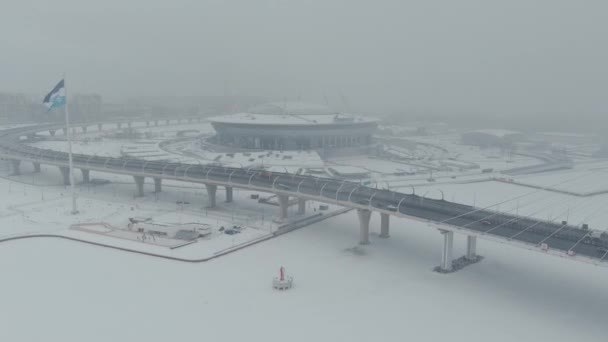 Rusia, San Petersburgo, 22 de enero de 2022: El avión no tripulado vuela a lo largo del puente de cable hacia el nuevo estadio Gazprom Arena en una tormenta de nieve, la bandera del equipo Zenit se balancea sobre un enorme asta de la bandera — Vídeo de stock