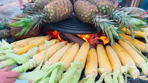На большом гриле, свежая кукуруза и ананасы жарят на открытом воздухе, кукурузные початки крутятся даже для приготовления пищи, праздник для большого количества людей — стоковое видео