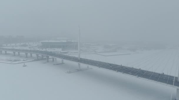 Russie, Saint-Pétersbourg, 22 janvier 2022 : Le drone vole le long du pont à haubans vers le nouveau stade Gazprom Arena dans une tempête de neige, le drapeau de l'équipe Zenit oscille sur un énorme mât de drapeau — Video