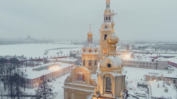 Imágenes aéreas de la fortaleza de Pedro y Pablo en una tormenta de nieve en una noche de invierno, iluminación nocturna, la catedral de Petropavlos brilla con luz dorada, vistas de San Petersburgo en el fondo — Vídeo de stock