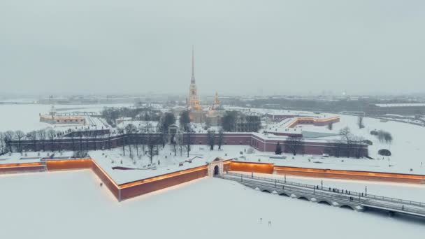 Aerial footage of the Peter and Paul Fortress in a snow storm in a winter evening, night illumination, Petropavlos Cathedral shines with golden light, sights of St. Petersburg in the background — Stock Video