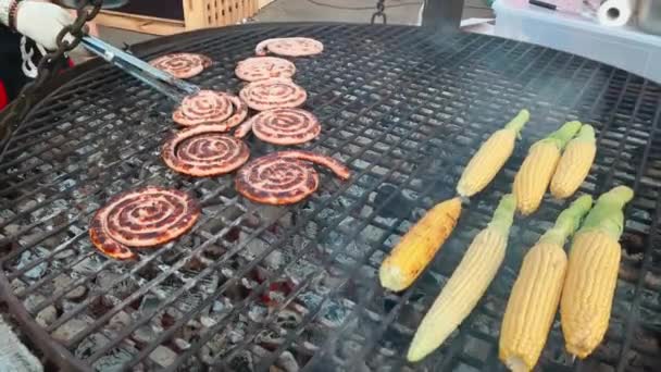 El chef con pinzas viendo la preparación de salchichas, maíz fresco y salchichas en espiral delgadas se fríen al aire libre, mazorcas de maíz, salchichas se hilan cada vez cocinar, un día de fiesta para un gran número de personas — Vídeo de stock