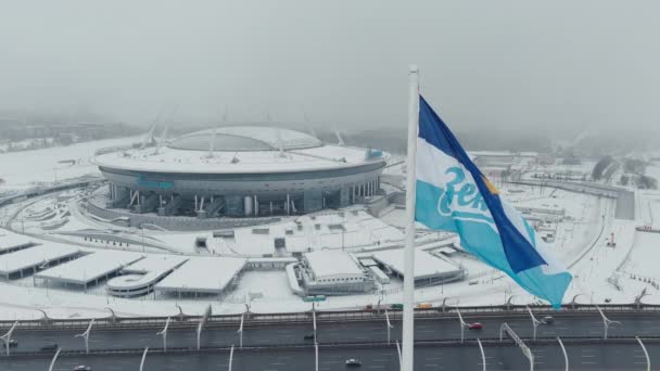 Russland, St. Petersburg, 22. Januar 2022: Die Drohne fliegt in der Nähe des neuen Stadions Gazprom Arena im Schneesturm, die Flagge des Zenit-Teams schwankt an einem riesigen Fahnenmast, viele Autos auf der Autobahn — Stockvideo