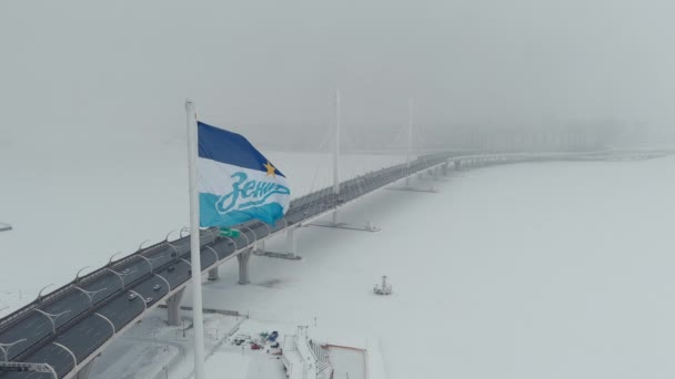 러시아, 상트페테르부르크, 2022 년 1 월 22 일:제 닛 축구 팀 깃발 이 눈보라 속에서 거대 한 깃대 위를 날아가고, 드론 이 새 경기장 가즈 프롬 아레나 근처의 케이블에 그대로 남아 있는 다리를 따라 날아가다 — 비디오