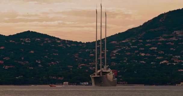 Obrovská jachta konceptuálního designu se třemi stěžni stojí v zátoce St. Tropez při západu slunce, jachta točí kolem kotvy, malé čluny plachty téměř, svah v pozadí, růžová obloha — Stock video
