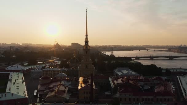 Le drone vole vers l'ange d'or sur la croix de la forteresse Pierre et Paul au lever du soleil, reflet du ciel orange sur l'eau, ponts-levis Troitsky et Liteiny — Video