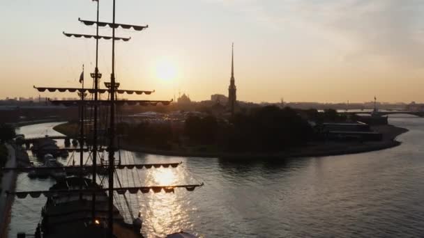 Drónok repülnek keresztül egy régi fregatt árbocain a Neva folyó felett a Péter és Pál erődbe napkeltekor, tükrözve a narancssárga eget a vízen. — Stock videók