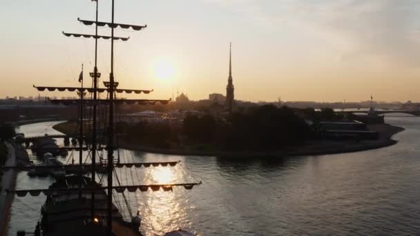 Drone vuelo a través de los mástiles de una antigua fragata sobre el río Neva a la fortaleza de Pedro y Pablo al amanecer, reflejo del cielo naranja en el agua — Vídeo de stock