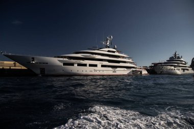 Günbatımında Monaco limanında iki büyük yat, motorlu teknenin parlak tahtaları, krom kaplama tırabzanlar, marinada demirli megayacht, parlak tahtada güneş yansıması, arka planda dağ