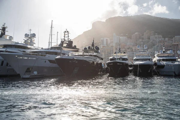 Una gran cantidad de enormes yates en el puerto de Mónaco al atardecer, la montaña está en el fondo, tablero brillante de la lancha a motor, megayates están amarrados en el puerto deportivo, reflejo del sol en tablero brillante, contraluz — Foto de Stock