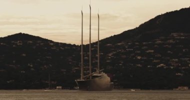 Günbatımında St. Tropez körfezinde üç direkli büyük bir kavramsal tasarım yatı duruyor. Yat çapasının etrafında dönüyor. Küçük tekneler neredeyse yelken açıyor.