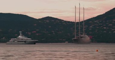 Fransa, St. Tropez, üç direkli ve motorlu büyük yatlı büyük bir konsept tasarım yatı gün batımında körfezde duruyor, yat çapasının etrafında dönüyor, arka planda yamaç var.