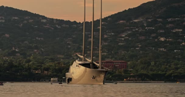 日没時にセント・トロペス湾に3つのマストを持つ概念的なデザインの巨大なセーリングヨット、ヨットはそのアンカーの周りを回転し、小さなボートはほぼ帆、背景の斜面、ピンクの空 — ストック動画