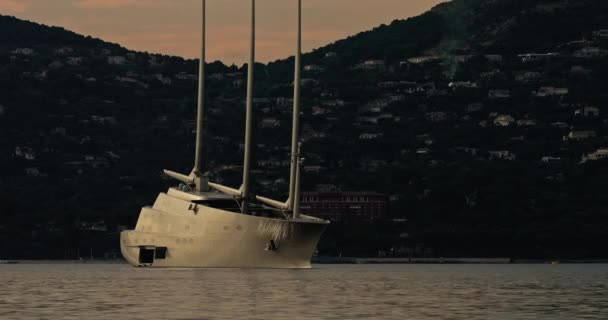 Огромная парусная яхта концептуального дизайна с тремя мачтами стоит в бухте Сан-Тропе на закате, яхта вращается вокруг своего якоря, маленькие лодки плывут почти, склон на заднем плане, розовое небо — стоковое видео