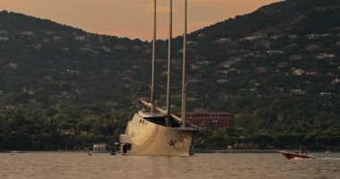 Fransa, St. Tropez, 31 Mayıs 2017: Günbatımında St. Tropez körfezinde üç direkli büyük bir yelkenli yatının hızlandırılması, yat çapasının etrafında dönüyor, küçük tekneler neredeyse yelken açıyor, bir yamaç.