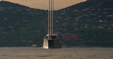 Günbatımında St. Tropez körfezinde üç direkli büyük bir kavramsal tasarım yatı duruyor. Yat çapasının etrafında dönüyor. Küçük tekneler neredeyse yelken açıyor.