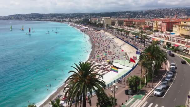 Muita gente na praia principal de Nice - França num dia de sol claro, água azul-turquesa, guarda-sóis do sol, uma atracção local - a Promenade des Anglais, barcos com uma vela no horizonte — Vídeo de Stock