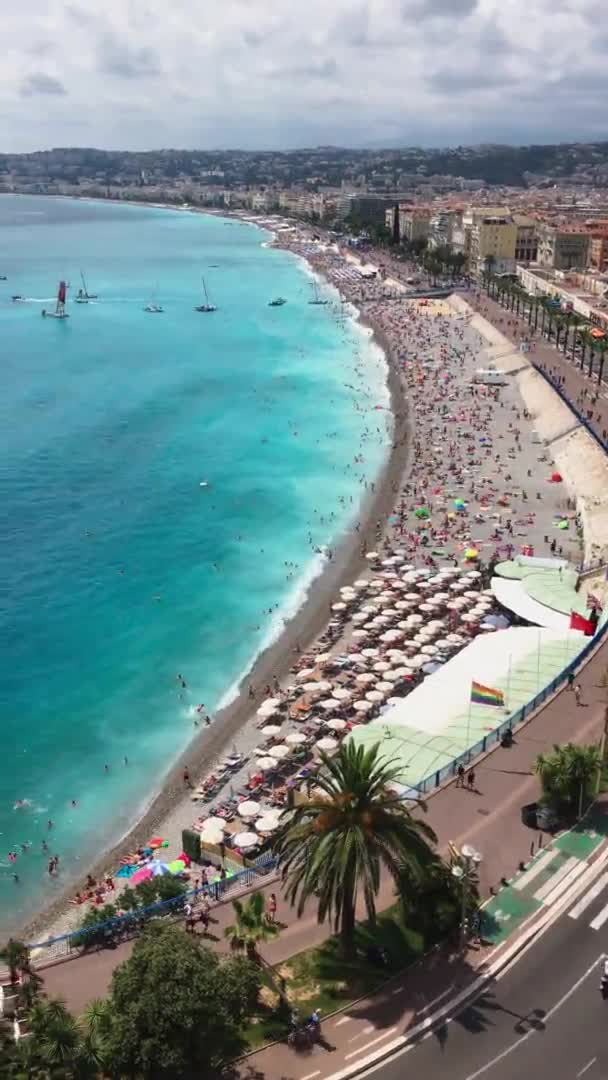 Muita gente na praia principal de Nice - França num dia de sol claro, água azul-turquesa, guarda-sóis do sol, uma atracção local - a Promenade des Anglais, barcos com uma vela no horizonte — Vídeo de Stock