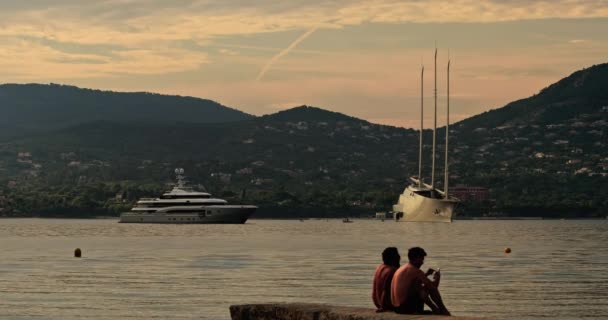 Frankrike, St Tropez, 31 Maj 2017: En stor segelbåt av konceptuell design med tre master och motor mega yacht vid solnedgången, ungdomar sitter på en klippa efter bad, lutning i bakgrunden — Stockvideo