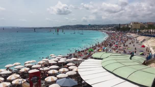 Un sacco di gente sulla spiaggia principale di Nizza - Francia in una chiara giornata di sole, acqua turchese, ombrelloni dal sole, un'attrazione locale - la Promenade des Anglais, barche con una vela all'orizzonte — Video Stock