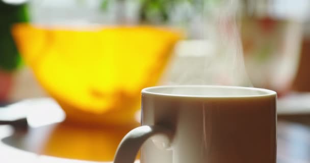 Ранок чашки кави або чаю в сонячний день чекає на столі, є пара, яка освітлюється сонцем з вікна, яскравий кольоровий фон, затишна кухня — стокове відео