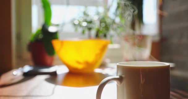 Poranna filiżanka kawy lub herbaty w słoneczny dzień czeka na stole, jest para, która jest oświetlona przez słońce z okna, jasne tło koloru, przytulna kuchnia — Wideo stockowe