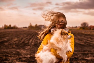 Rüzgarlı, sıkıcı bir sonbahar günü. Pomeranya köpeğiyle beyaz bir kızın açık hava portresi. Şiddetli rüzgarda esen uzun sarı saçlar. Fırtına. Arkaplan olarak ülke alanları manzarası.