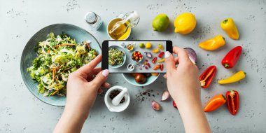 Kadın taze, baharlı, akıllı telefonlu sebze salatası malzemelerinin fotoğrafını çekiyor. Yemek yaparken telefon kullanan bir blogcu. Yaşam tarzı eğilimi - sosyal medyada yiyecek resimlerinin paylaşımı ve paylaşımı.