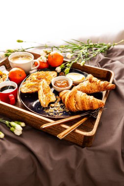 Yatakta, kırsal ahşap tepside Avrupalı kahvaltı. Kahve, kruvasan, reçel, tereyağı, meyve ve çiçek. Bir çift için tembel, romantik bir sabah yemeği. Poster düzeni, ücretsiz kopyalama alanı.
