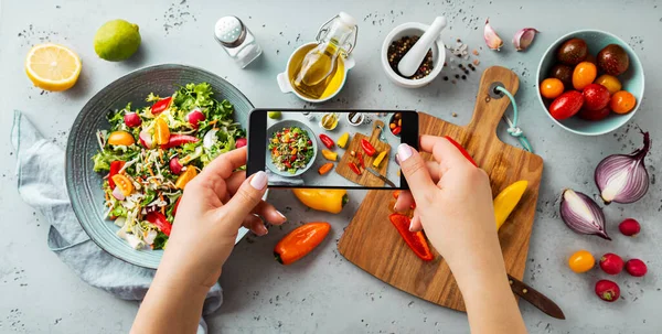 Woman taking photo of fresh, spring, vegetable salad ingredients with smartphone. Blogger using phone while cooking. Lifestyle trend - posting and sharing food pictures (images) on social media.