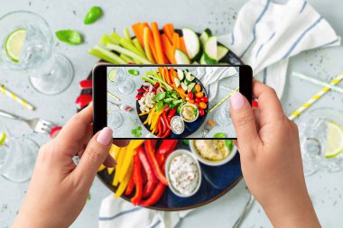 Tabakta taze, çiğ ve dilimlenmiş sebzelerin akıllı telefonuyla fotoğrafını çeken bir kadın. Yemek blogcusu yemek yakalamak için telefon kullanıyor. Yaşam tarzı eğilimi - sosyal medyada yiyecek resimlerinin paylaşımı ve paylaşımı.