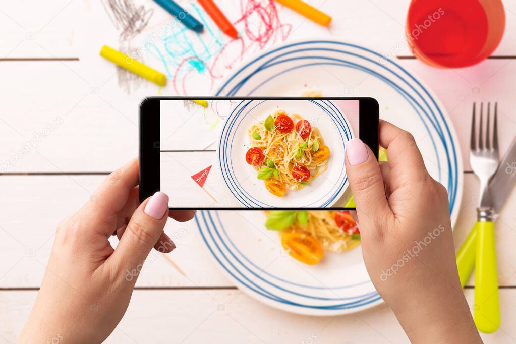 Woman taking photo of kid's dinner (spaghetti) with smartphone. Food blogger using phone to capture meal. Lifestyle trend - posting and sharing food pictures on social media.