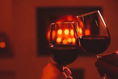 Romantik bir buluşma. İki kadeh kırmızı şarapla kadeh kaldıran bir çift. Arkaplan olarak şömine - ücretsiz kopyalama (metin) alanı. Noel ya da Sevgililer Günü havası.