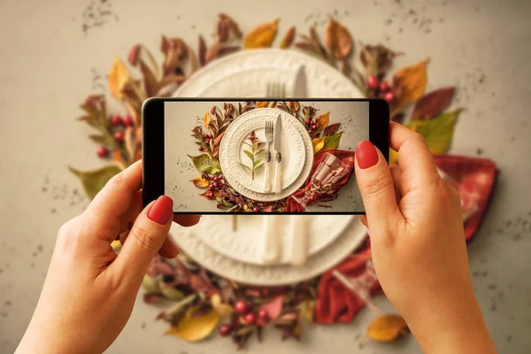 Woman taking photo of autumn (fall) table setting design idea with smartphone. Blogger, influencer or stylist capturing Thanksgiving home decorations for social media.