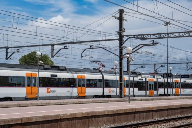 Vilanova i la Geltru, İspanya: Katalan bölgesel demiryolu sisteminin istasyondaki treni. Arka planda bulutlu mavi gökyüzüne karşı tren kabloları. RENFE banliyö treni. Barselona banliyö demiryolu hizmeti.